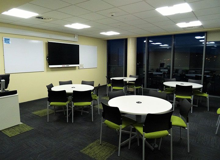 A photo of a classroom with four circular tables each surrounded with six green chairs with the lights on