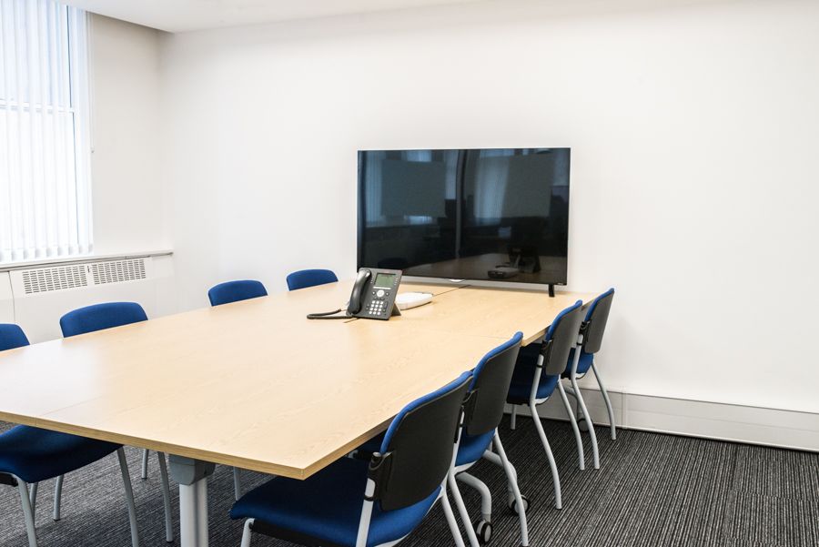 A photo of inside a meeting room with a large TV, a telephon and eight blue chairs around a table